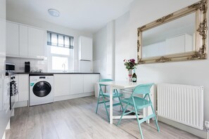 Een moderne keuken met witte kasten, een wasmachine, een eettafel met turquoise stoelen, en een grote spiegel aan de muur.