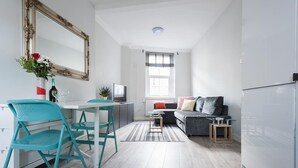 Une cuisine moderne avec des meubles blancs, un coin repas avec des chaises de couleur turquoise, un canapé gris et un grand miroir.