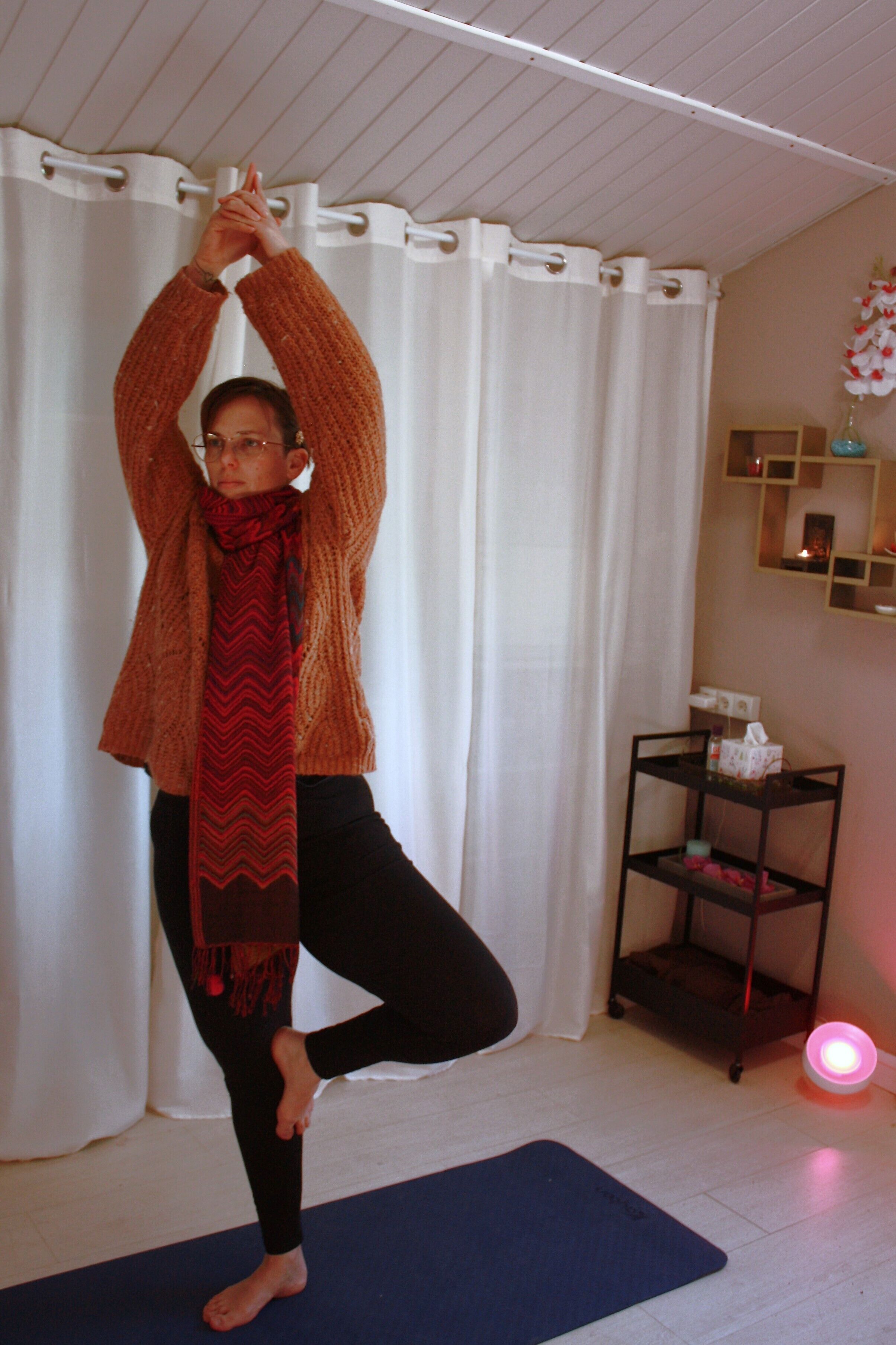 A person performing a yoga pose in a room with a curtain, a small shelf with items, and a yoga mat on the floor.