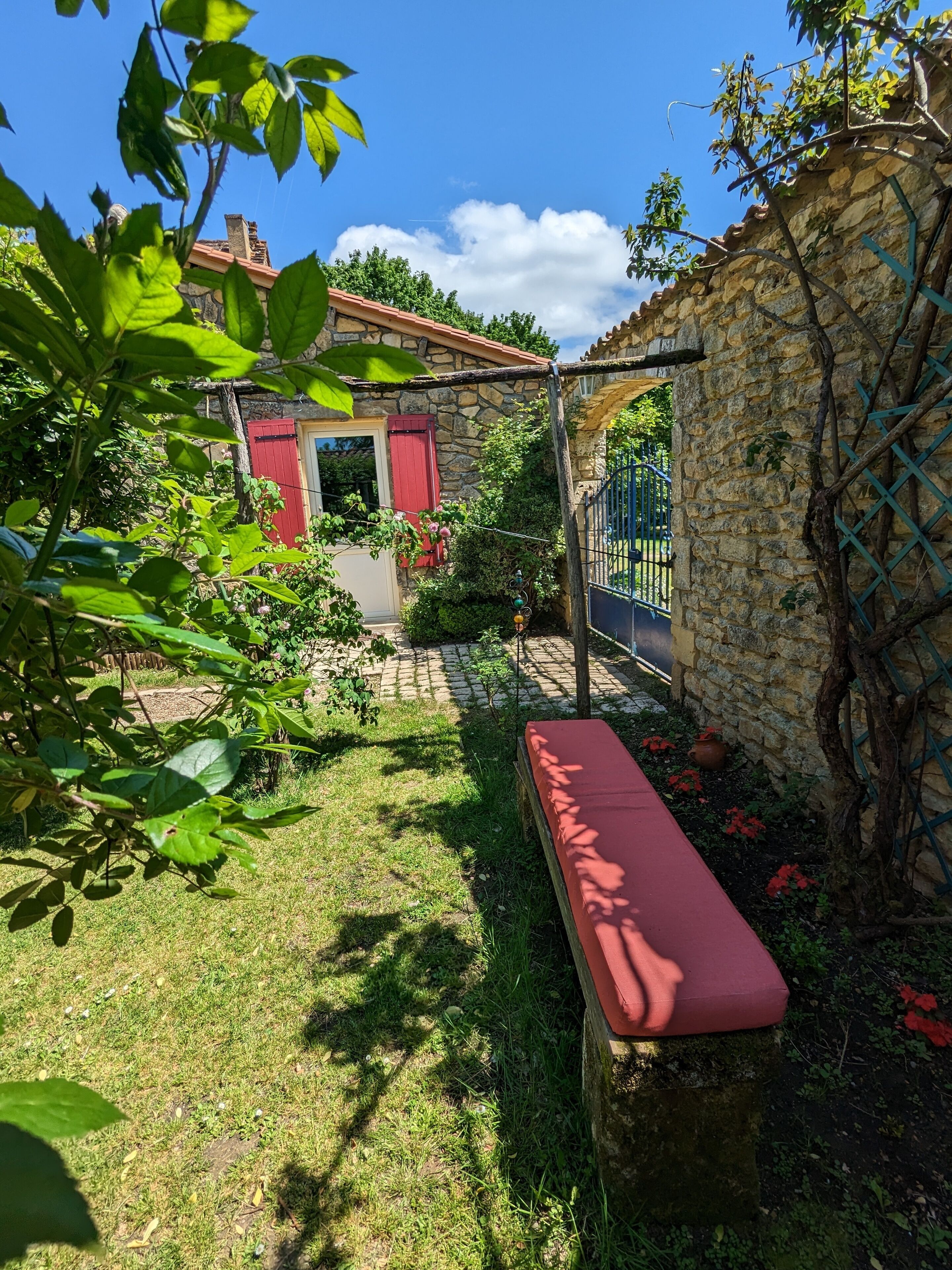 Uma casa de pedra com porta e janelas em vermelho, um muro de pedra com plantas trepadeiras, um portão de metal e um banco no quintal.