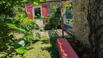 Uma casa de pedra com porta e janelas em vermelho, um muro de pedra com plantas trepadeiras, um portão de metal e um banco no quintal.