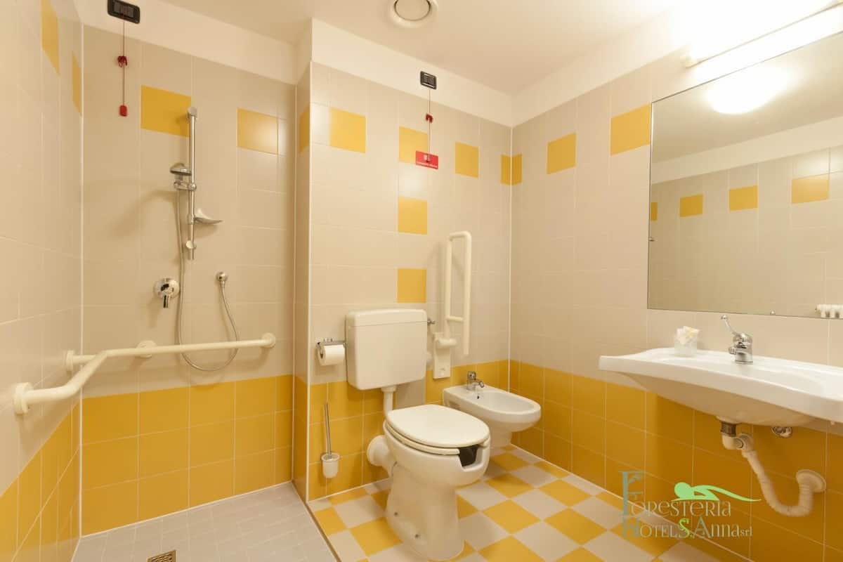 Un baño con azulejos amarillos y blancos, ducha, inodoro, bidé y lavamanos.