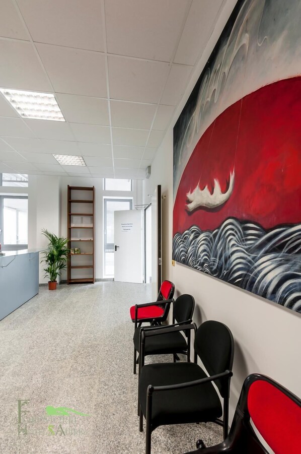 A reception area with a modern design, featuring a reception desk, a seating area with chairs, a bookshelf, and a large abstract painting on the wall. - Foresteria Sant'Anna (Ferrara)