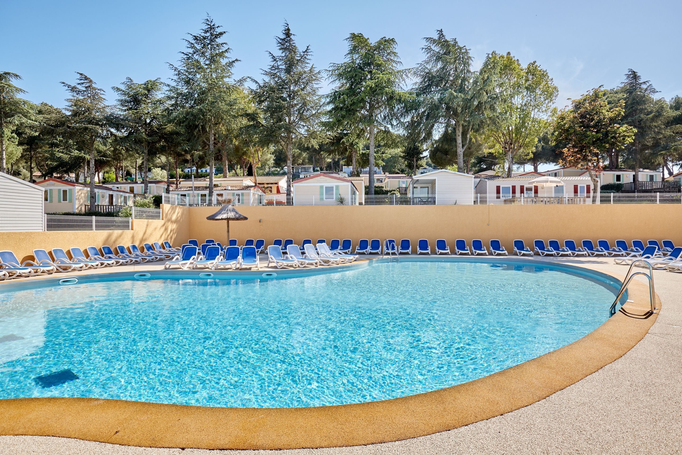 A swimming pool with blue chairs and a sandy deck, surrounded by trees and mobile homes.