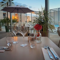 Mesa lista para comer, con copas de vidrio transparente, un jarrón con una flor roja y vista a un edificio con fachada decorativa.