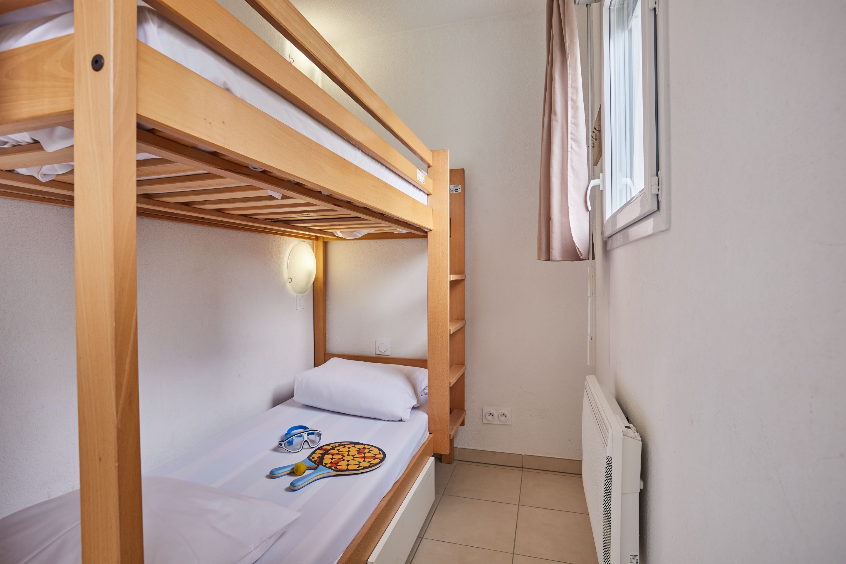 Una cama individual con una raqueta de tenis y gafas de sol, una ventana con cortinas y un radiador en la pared.