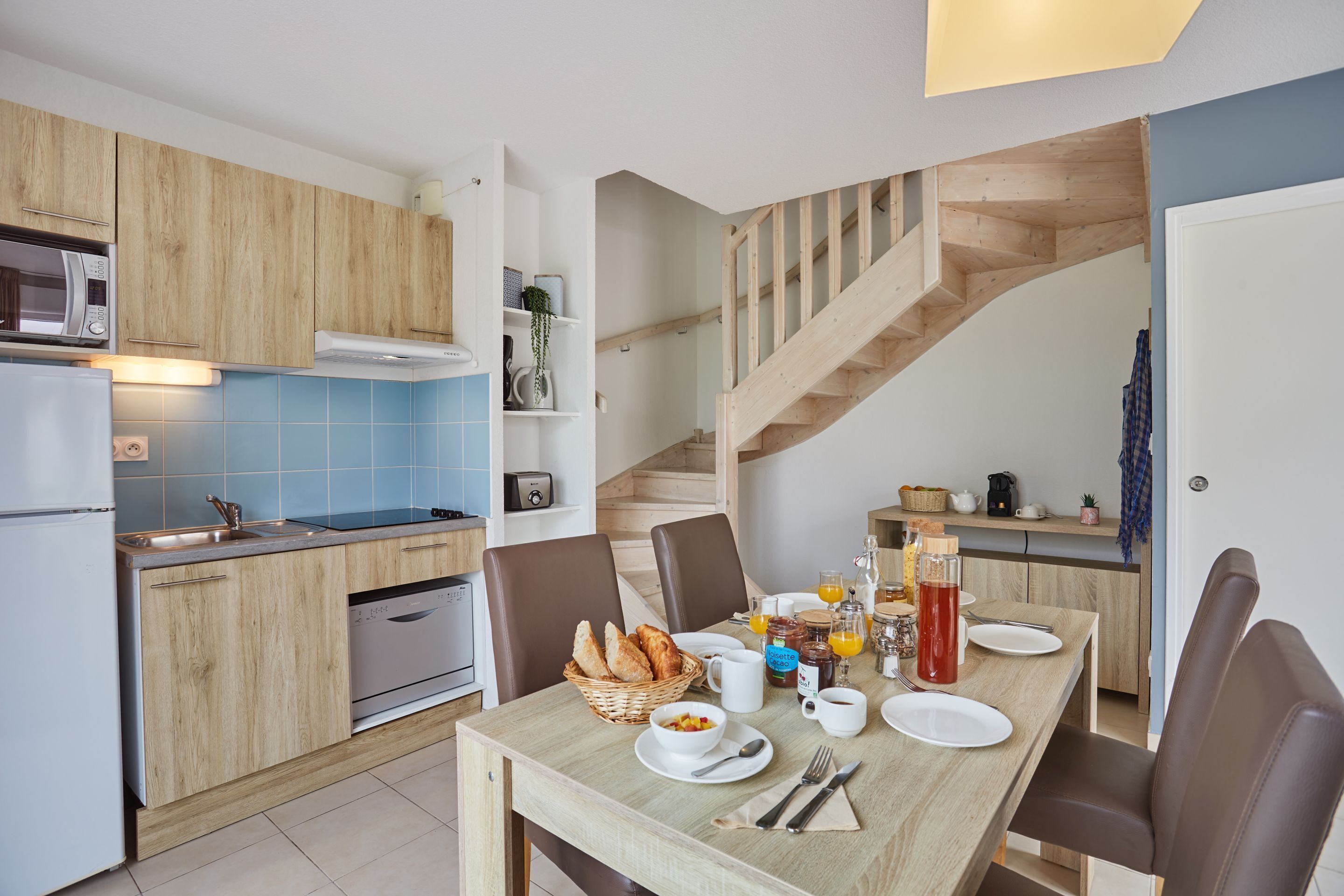 A modern kitchen with wooden cabinets, a dining table set for breakfast, and a wooden staircase.