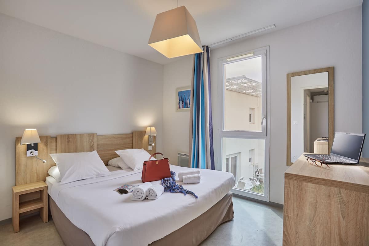 Ett hotellrum med en säng, sängbord, ett skrivbord med en bärbar dator och ett fönster med gardiner.