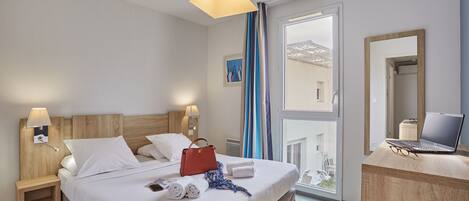 A hotel room with a bed, bedside tables, a desk with a laptop, and a window with curtains.