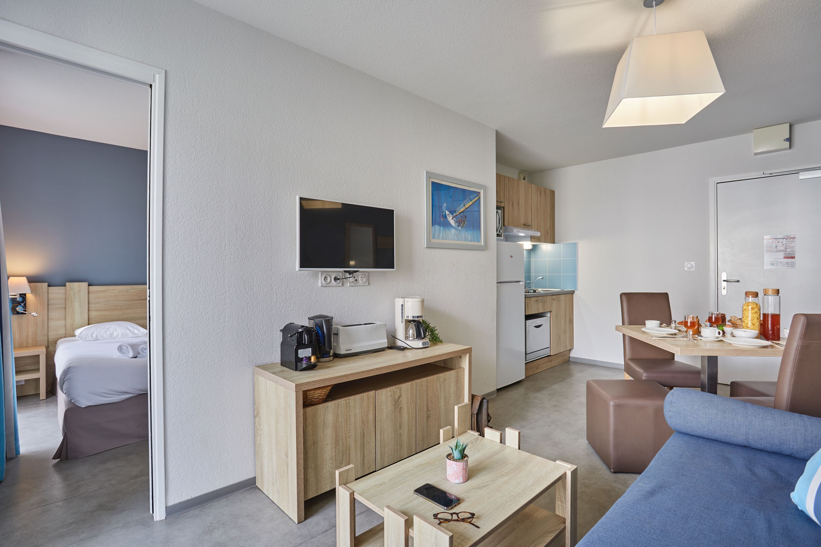 A modern hotel room with a bed, a TV, a kitchenette, a dining area, and a living space.