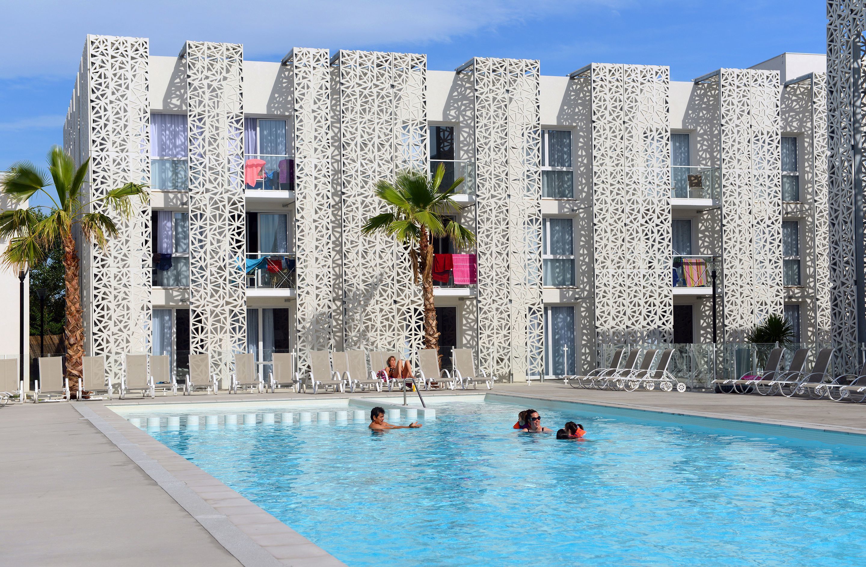 A pool area with lounge chairs and palm trees, surrounded by a building with a geometric facade.