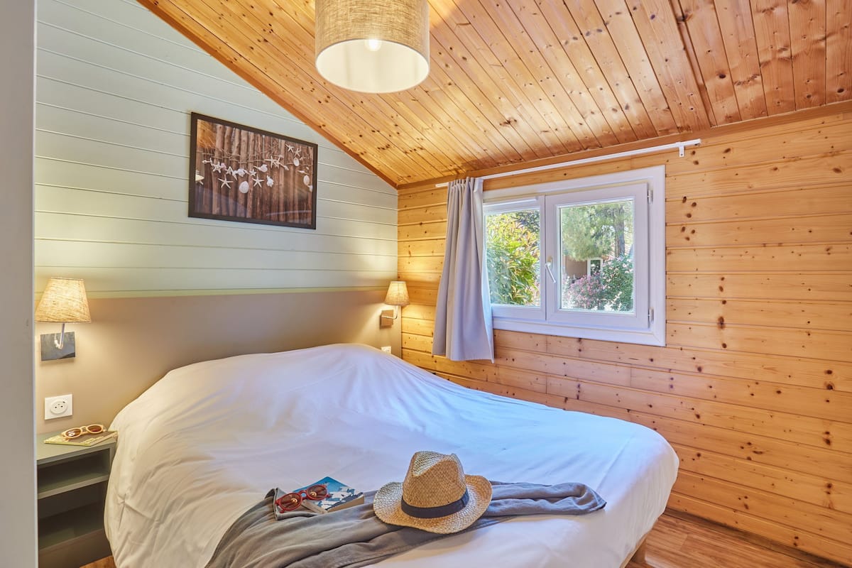 Una habitación acogedora con paredes de madera, una cama grande, dos lámparas de noche, un ventanal con cortinas y un cuadro colgado en la pared.