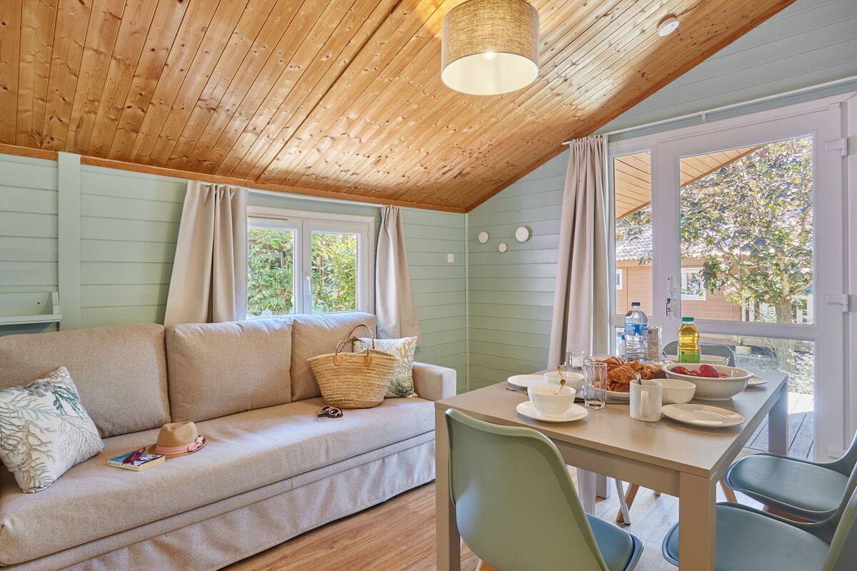 Un cuarto acogedor con techo de madera, un sofá con cojines, una mesa de comedor con sillas y vistas a zonas verdes.