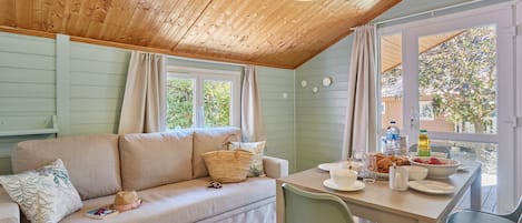 A cozy room with a wooden ceiling, a sofa with cushions, a dining table with chairs, and a view of greenery outside.