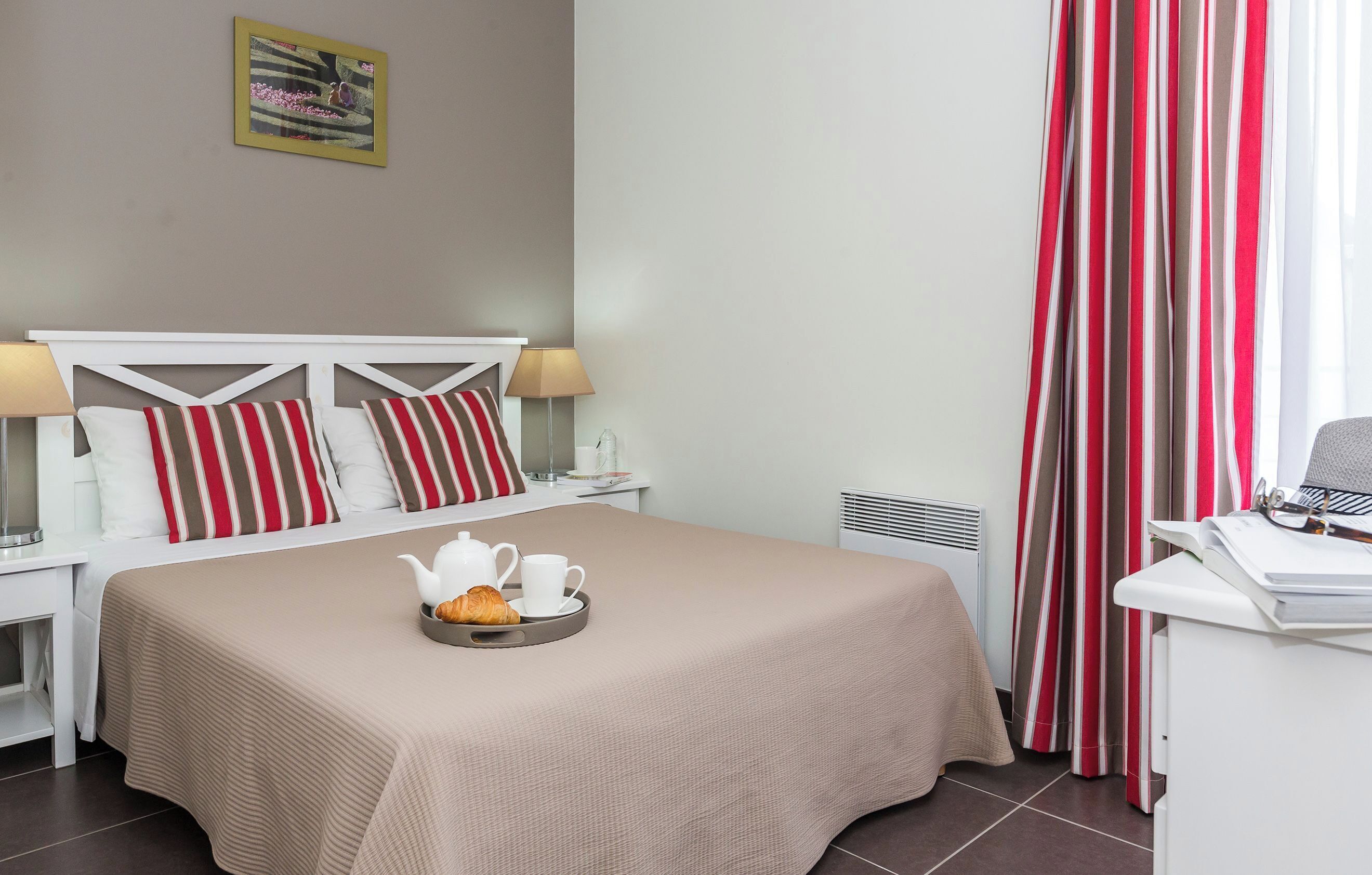 Een net opgemaakt bed met een dienblad croissants en een theepot, een nachtkastje met een lamp, en een bureau met een telefoon en papieren.