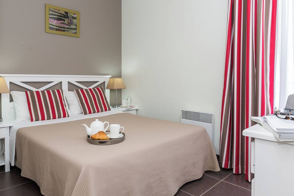 Een net opgemaakt bed met een dienblad croissants en een theepot, een nachtkastje met een lamp, en een bureau met een telefoon en papieren.