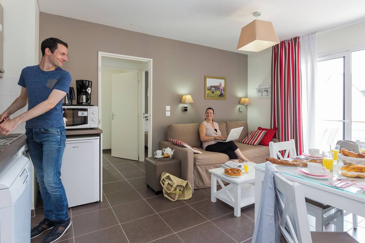 Een moderne woonkamer met een bank, een eettafel met ontbijtspullen, en een keukenhoek waar iemand eten bereidt.