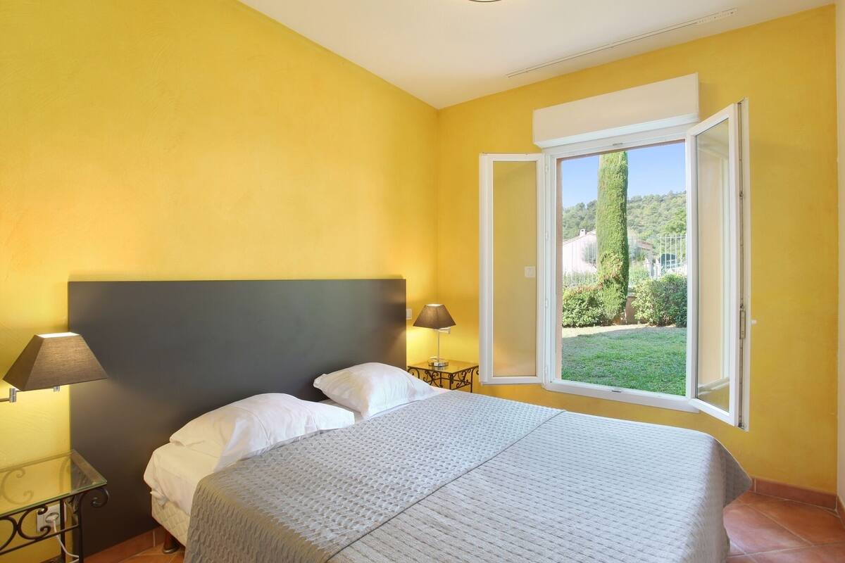 Un dormitorio con cama, dos lámparas de noche, un ventanal con persiana abierta y vistas a zonas verdes.