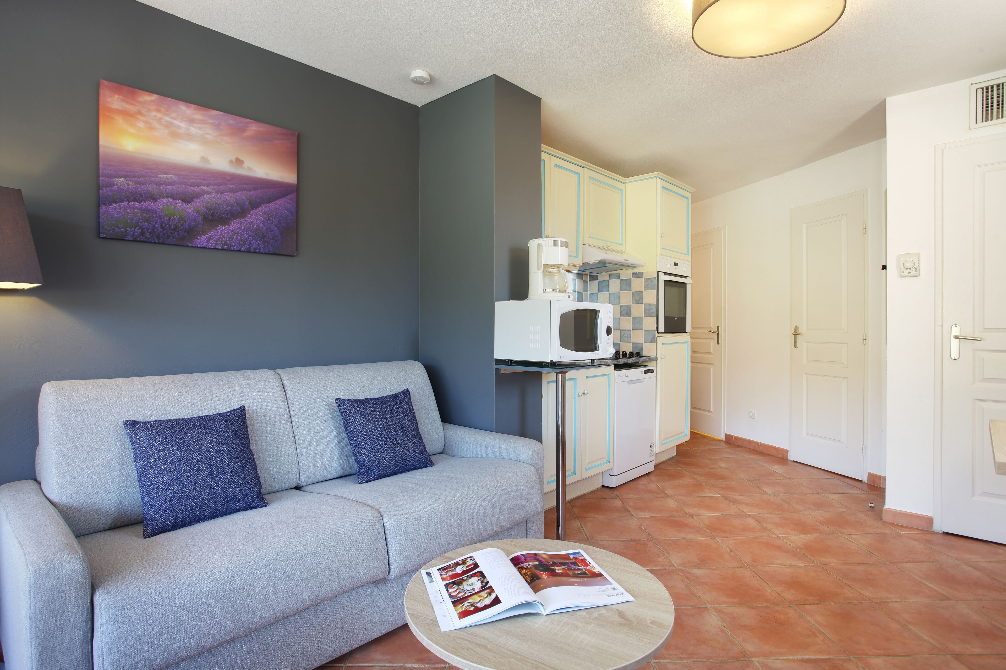 A living room with a sofa, a round coffee table with a magazine, a kitchen area with a microwave, and a door.