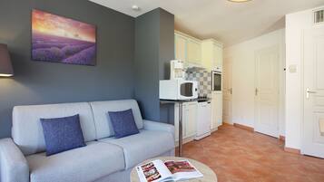 A living room with a sofa, a round coffee table with a magazine, a kitchen area with a microwave, and a door.