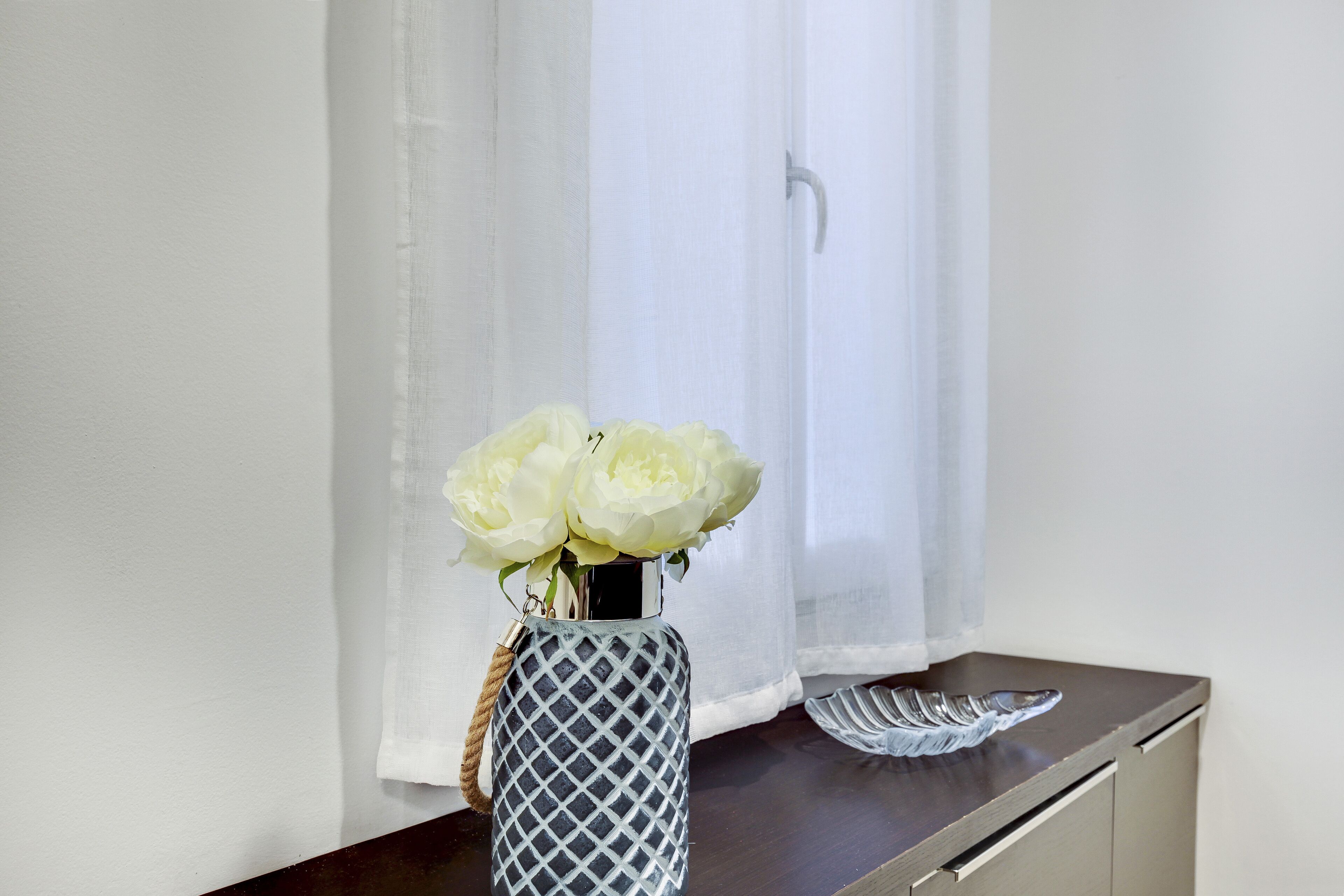 A vase with white flowers on a wooden surface near a window with sheer curtains.