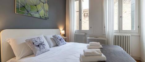 A neatly made bed with white linens and decorative pillows, a radiator, and a window with curtains.