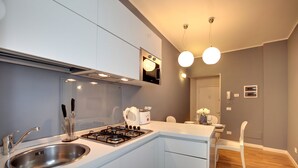 Una cocina moderna con un lavamanos de acero inoxidable, una estufa a gas y armarios blancos. Hay una zona de comedor con una silla blanca y una mesita.