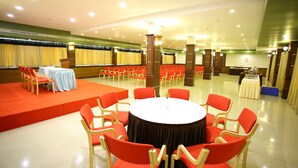 Un salón de conferencias con una mesa redonda rodeada de sillas rojas, un escenario con mantel azul y un fondo con cortinas.