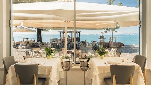 An indoor dining area with tables set for two, overlooking a beach and the sea. - Hotel Giardinetto (Garda)