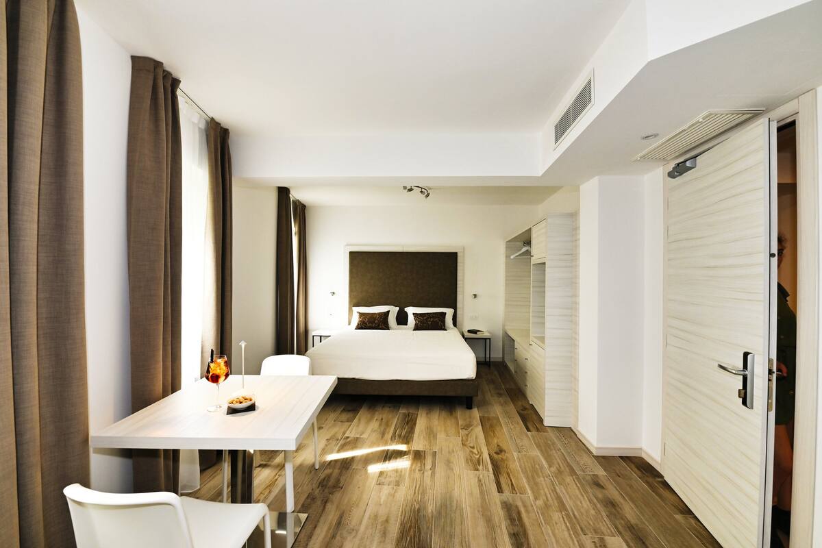 A modern hotel room with a large bed, a bedside table, a chair, and a view of the hallway.