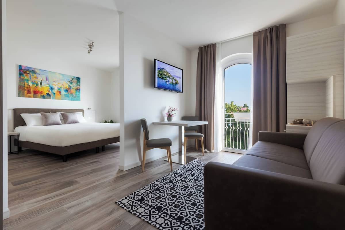 A modern hotel room with a bed, a sofa, a dining area, and a balcony with a view.