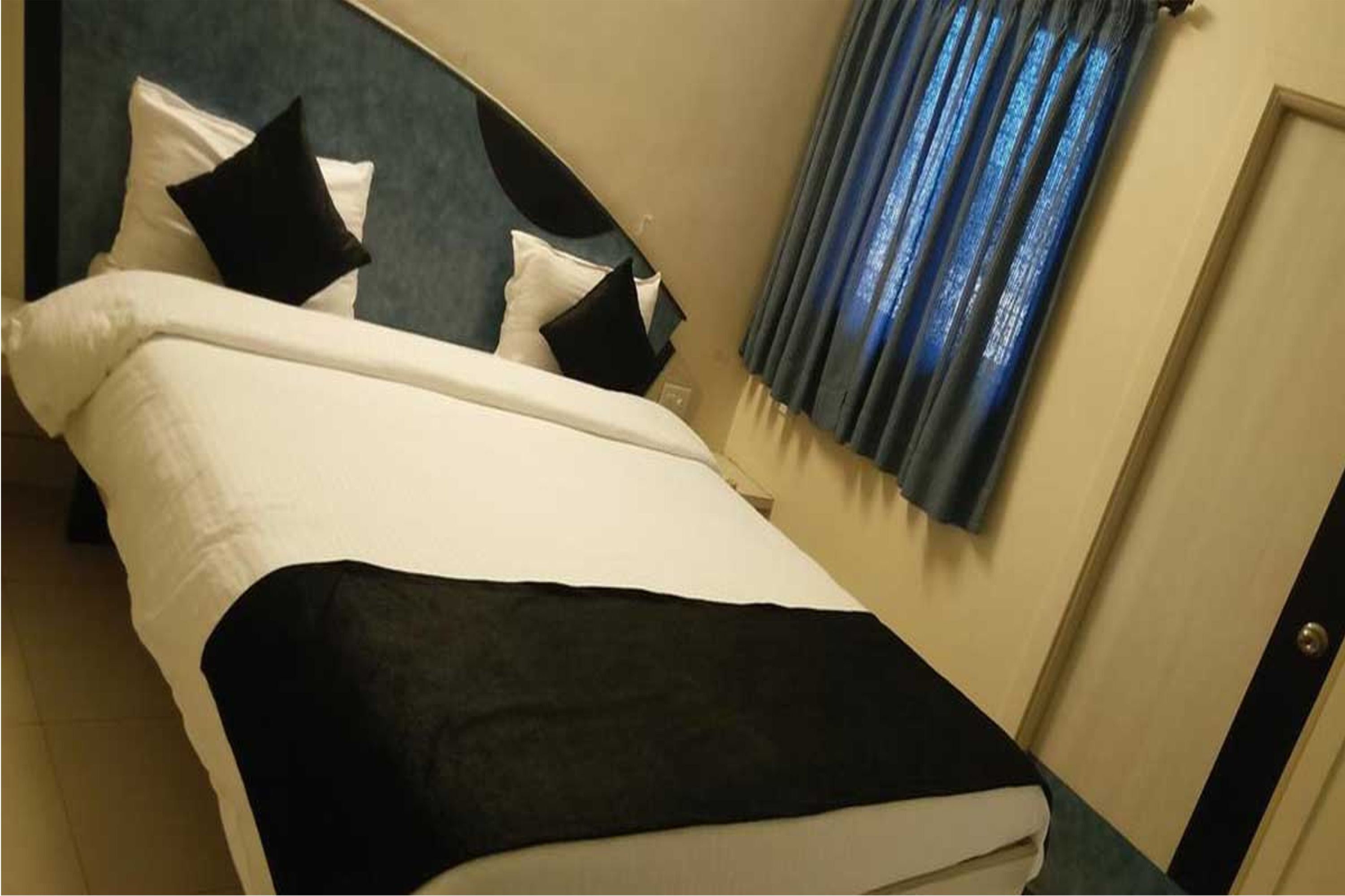 Una habitación de hotel con una cama, almohadas, un plaid negro y una ventana con cortinas azules.