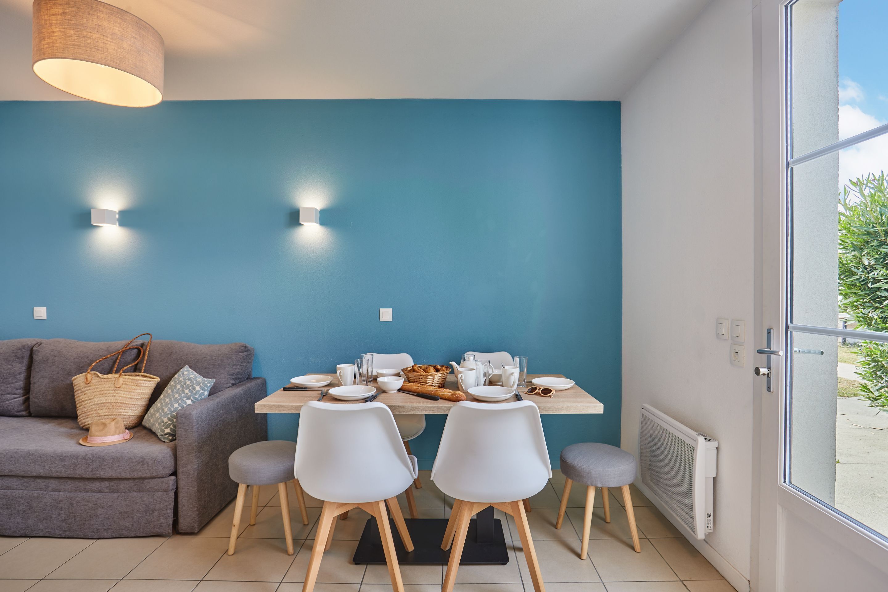 A modern living room with a blue accent wall, a grey sofa, a dining table set with chairs, and a large window with a view of greenery.