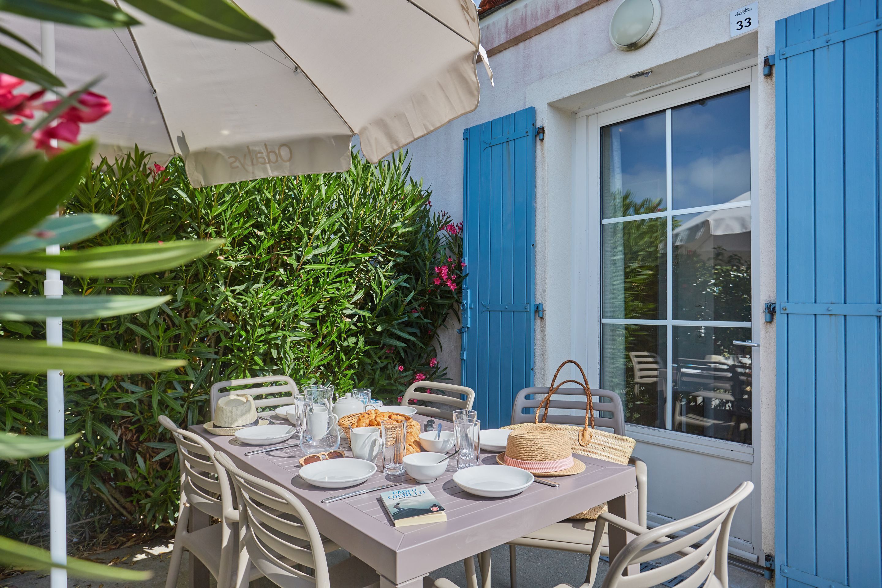 An outdoor dining area with a table set for four, surrounded by white chairs. There is a large umbrella providing shade, and the setting is next to a building with blue shutters and a window.