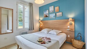 Un lit bien fait, avec une couette blanche et des oreillers, une lampe de chevet et une étagère fixée au mur, sur laquelle se trouvent un livre et un chapeau.