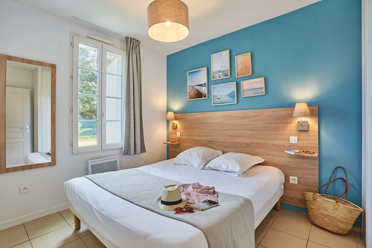 Un lit bien fait, avec une couette blanche et des oreillers, une lampe de chevet et une étagère fixée au mur, sur laquelle se trouvent un livre et un chapeau.