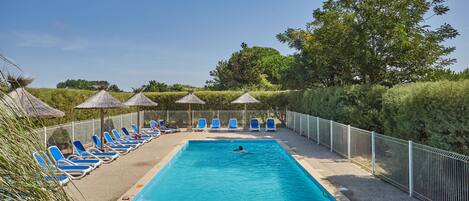 A rectangular swimming pool with blue water, surrounded by a white fence. There are blue lounge chairs and thatched umbrellas along the poolside.