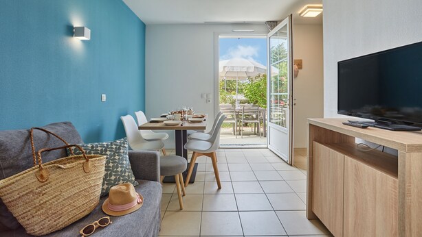 Un salon moderne comprenant un canapé, une table à manger avec des chaises, un téléviseur posé sur un meuble en bois et une grande fenêtre donnant sur l’extérieur.