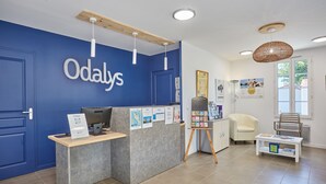 Un espace d’accueil avec un mur bleu, un comptoir de réception, un ordinateur, un coin salon avec une chaise et une petite table, et un présentoir à affiches.