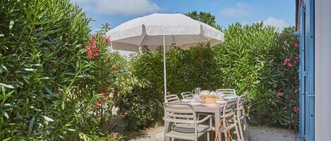 Ein Essbereich im Freien mit Tisch und Stühlen unter einem weißen Sonnenschirm, umgeben von üppigem Grün und rosa Blumen.