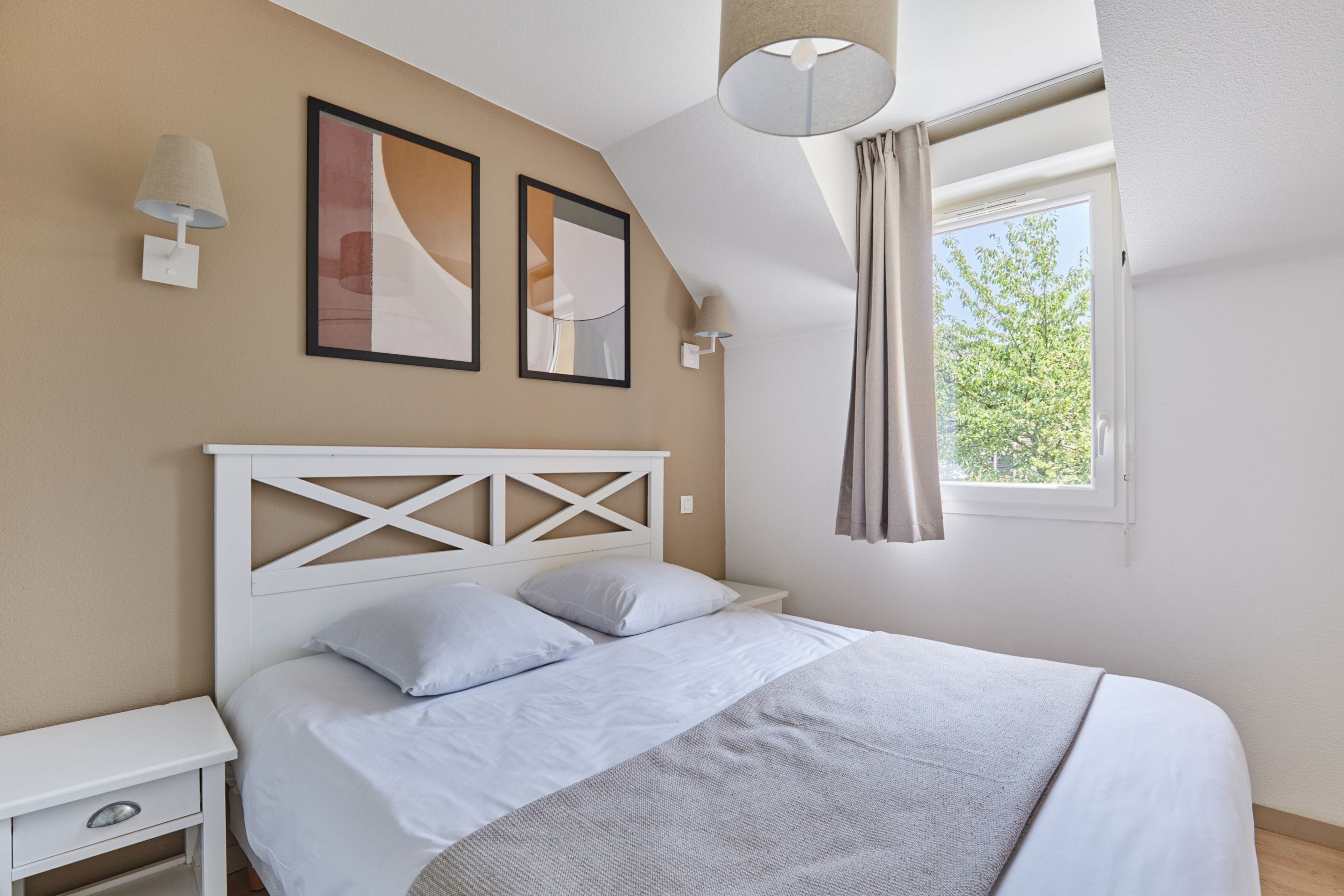 A bedroom with a bed, a nightstand, a wall-mounted lamp, and a window with curtains.