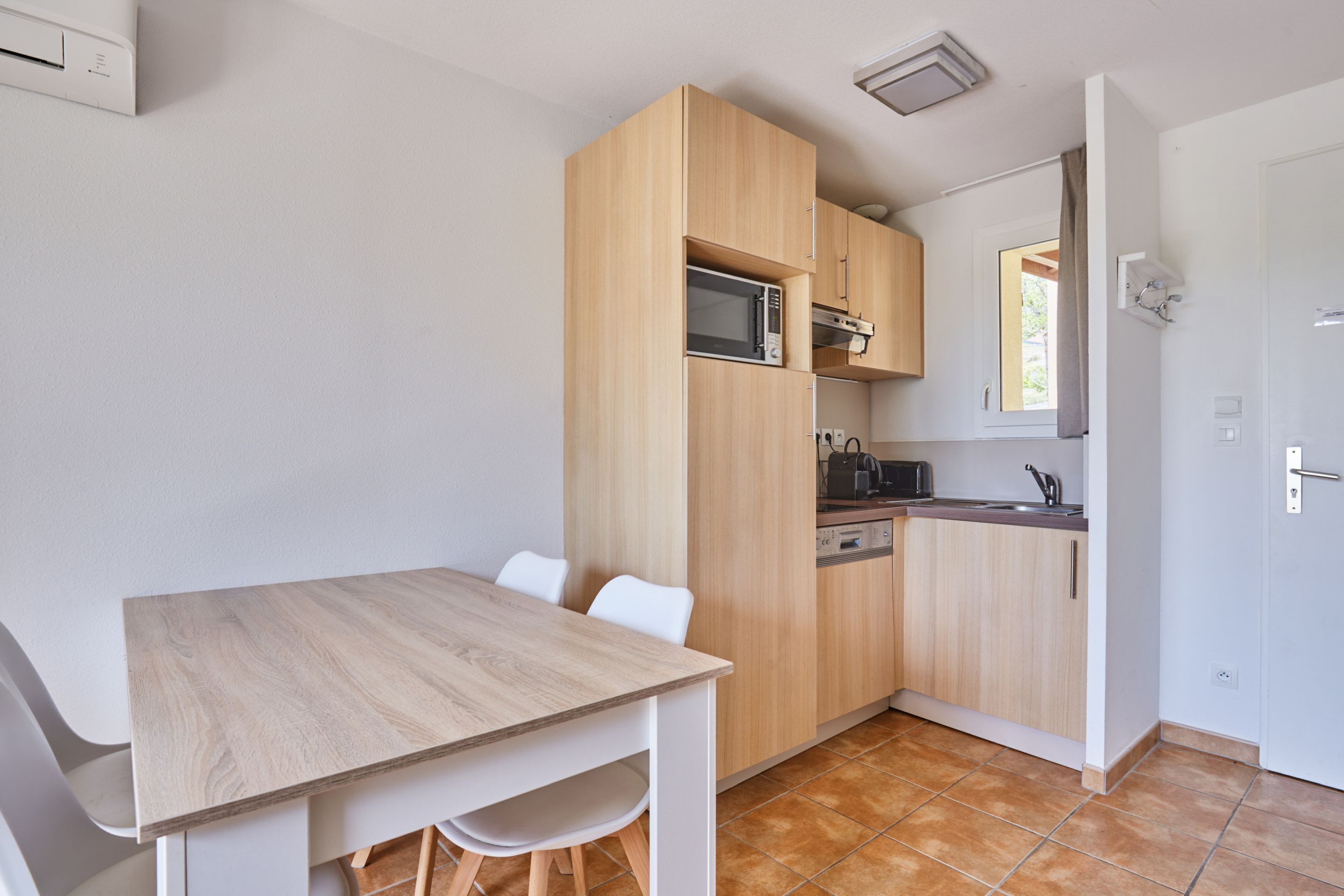 Een compacte keuken met houten kastjes, een magnetron en een spoelbak. Er is een eettafel met stoelen.