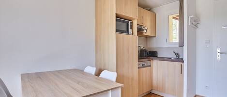 Een compacte keuken met houten kastjes, een magnetron en een spoelbak. Er is een eettafel met stoelen.