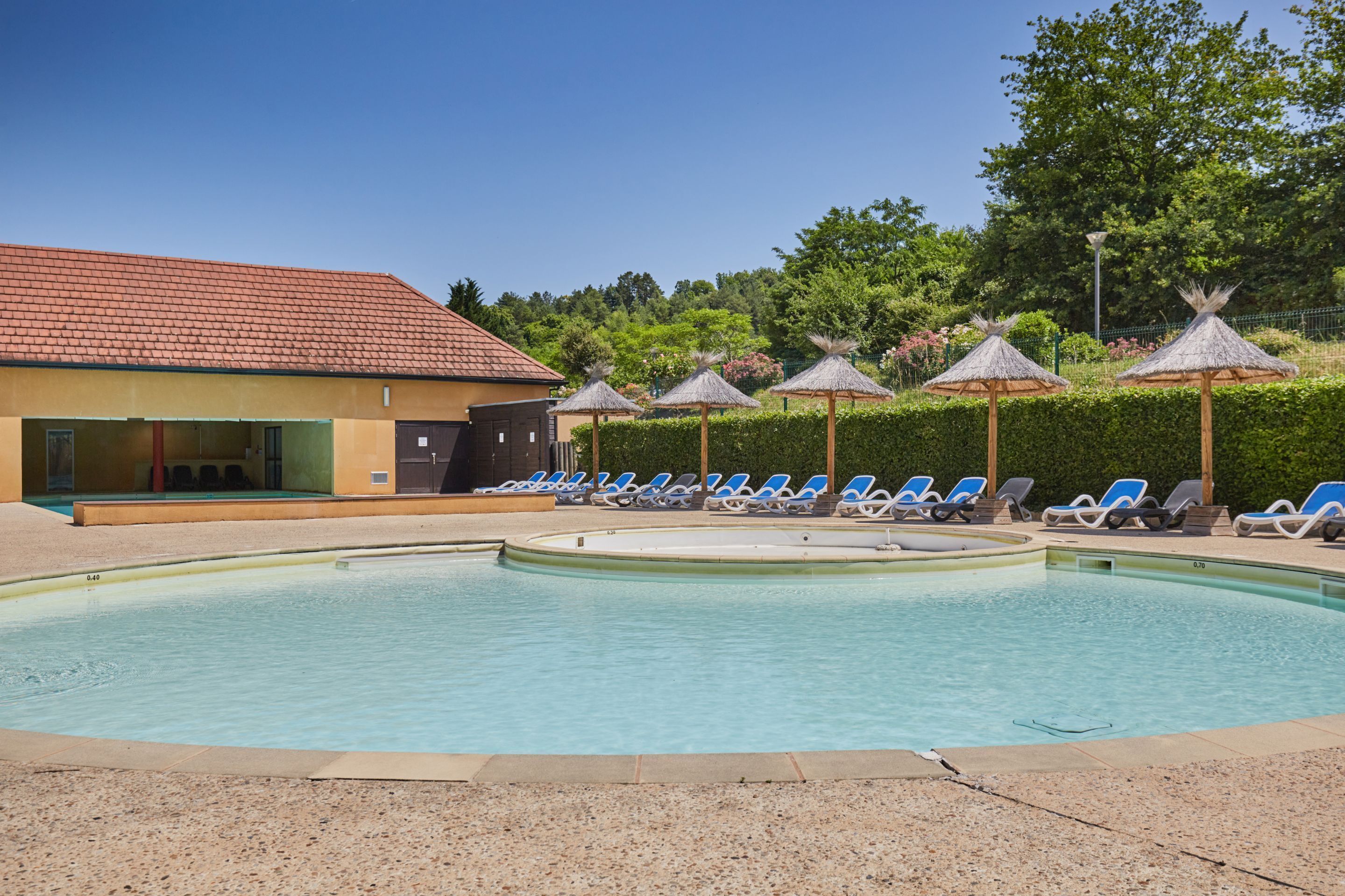 A swimming pool with lounge chairs and umbrellas, a building with a tiled roof, and a hedge in the background.