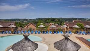 Un espace piscine agrémenté de chaises longues bleues et de parasols en chaume, entouré de bâtiments aux toits recouverts de tuiles rouges.
