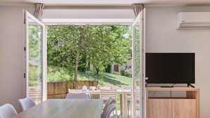Une chambre moderne équipée d’une table à manger, de chaises, d’un téléviseur et offrant une vue sur un jardin grâce à une porte ouverte.