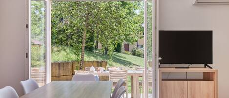 A modern room with a dining table, chairs, a TV, and a view of a garden through an open door.