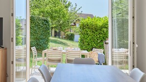 Un coin repas avec une table et des chaises, un téléviseur, et une vue sur un jardin et une maison, le tout visible à travers des portes vitrées.
