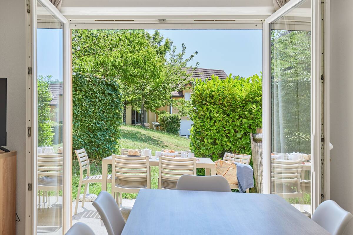 A dining area with a table and chairs, a television, and a view of a garden and house through glass doors.