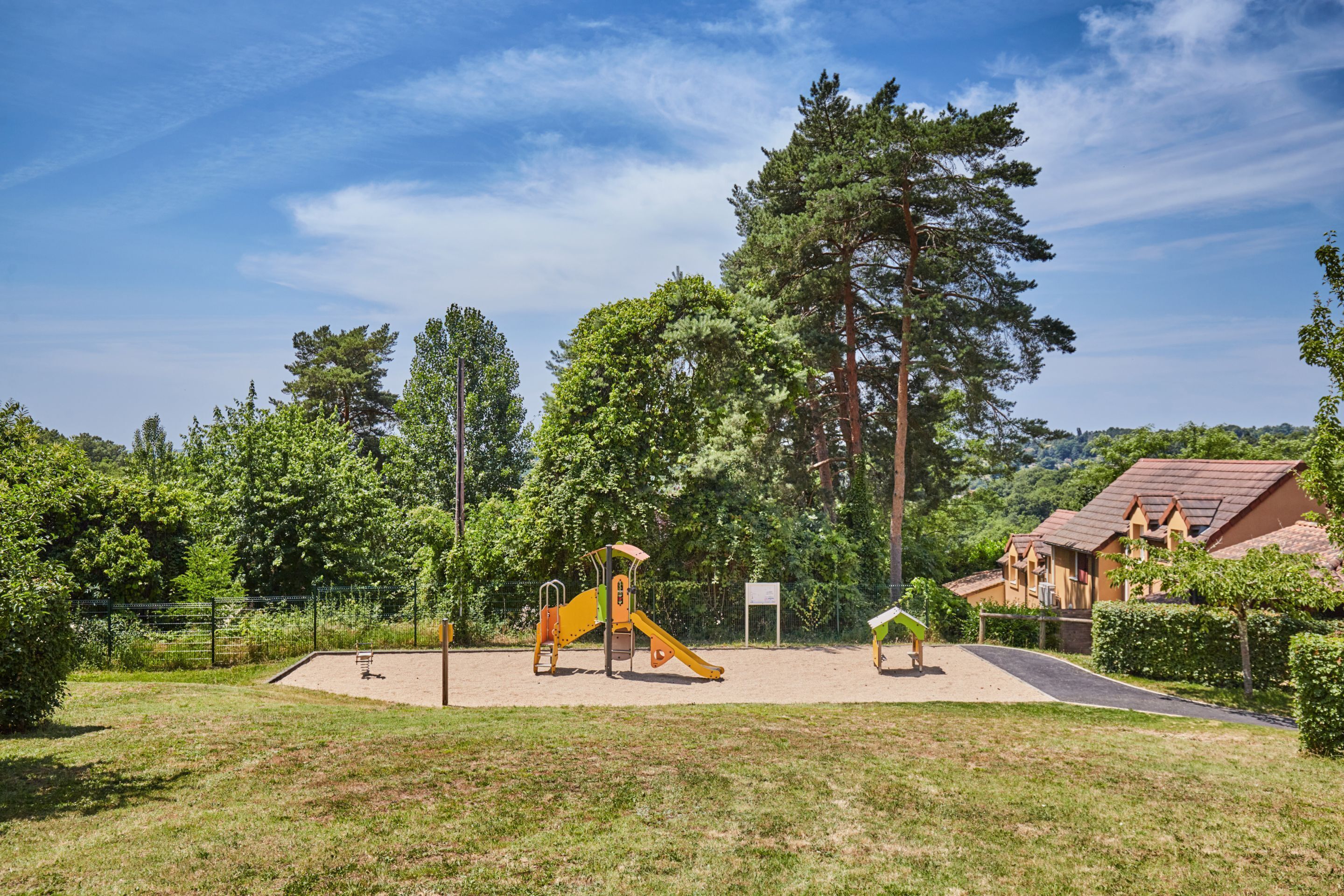 A playground with yellow slides and a fenced area, surrounded by trees and a building with a red roof.